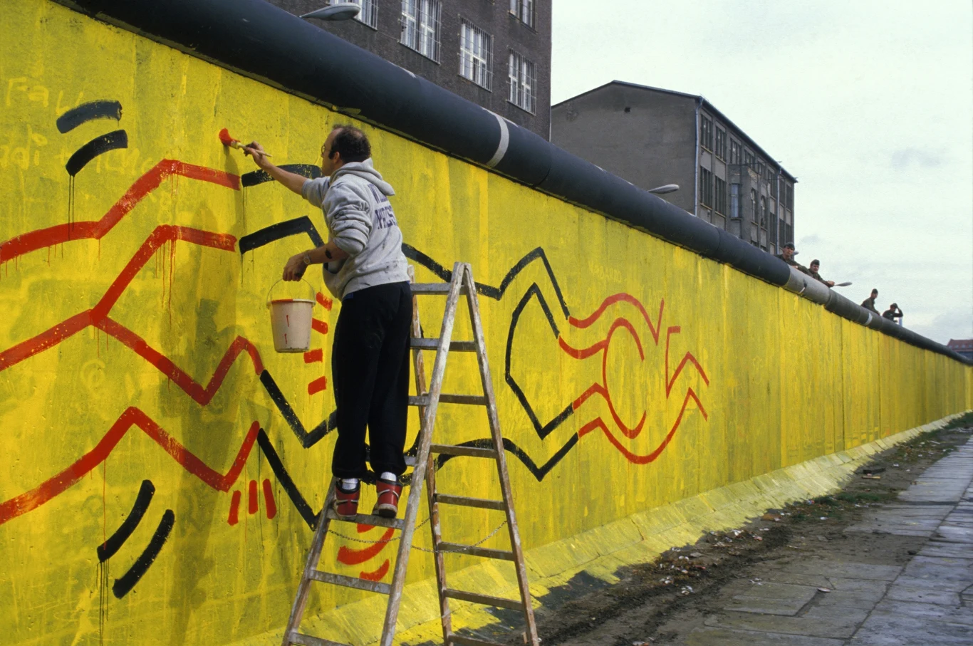 Mural Keithaa Heringa na murze berlińskim Mężczyzna stojący na drabinie maluje żywą żółto-czarną abstrakcyjną grafikę na długim fragmencie muru, w tle widać budynki industrialne i kilka osób idących wzdłuż ściany.