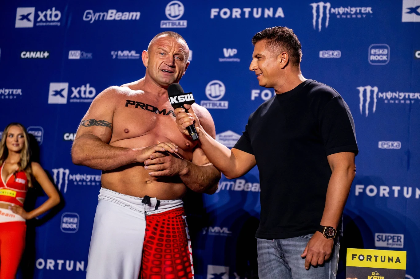 Mariusz Pudzianowski i Mateusz Borek przed galą KSW Dwóch mężczyzn na tle ściany z logotypami sponsorów, jeden z nich w stroju zawodnika MMA udziela wywiadu drugiemu trzymającemu mikrofon, w tle hostessa.