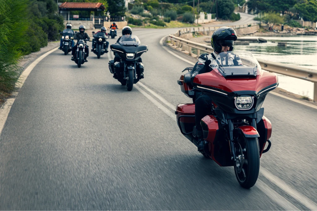 Grupa motocyklistów jadąca po asfaltowej drodze w pobliżu wody, prowadząca kolumnę osoba na czerwonym motocyklu, pozostali motocykliści na czarnych motocyklach podążają za nią w malowniczym otoczeniu drzew i zabudowań.