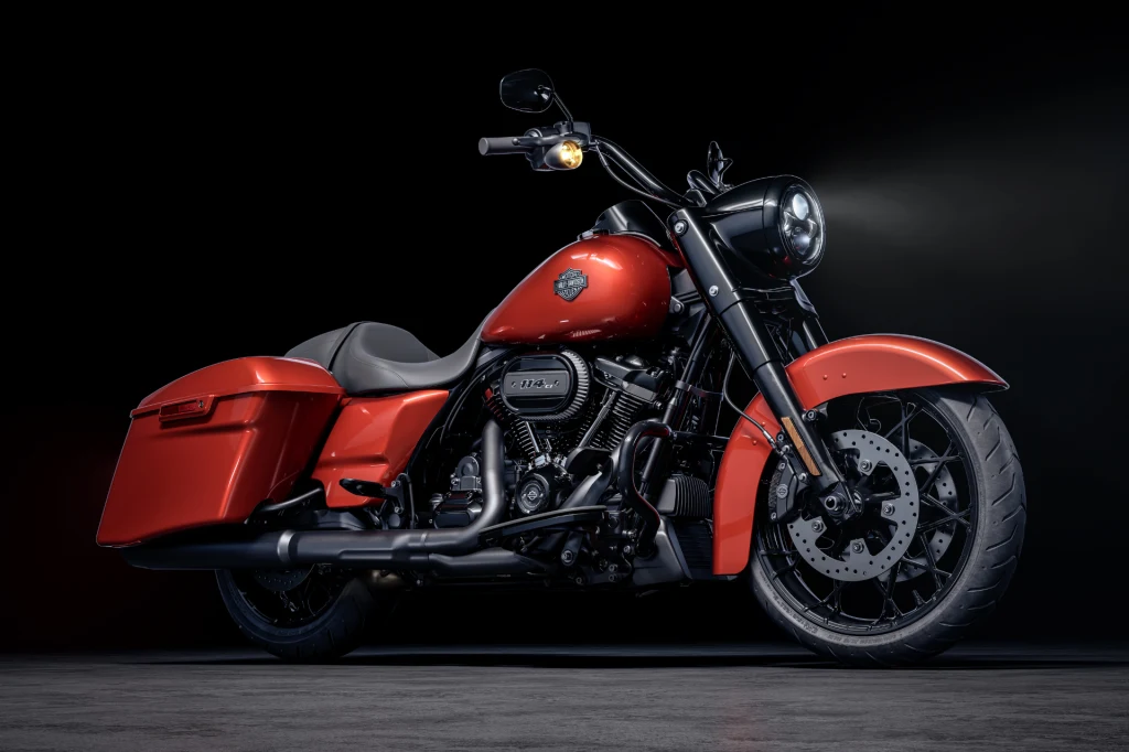 Czerwony motocykl Harley-Davidson z czarnymi detalami, stojący na tle ciemnego, minimalistycznego tła, wyróżniają się detale techniczne, masywne koła i charakterystyczne logo marki na baku.