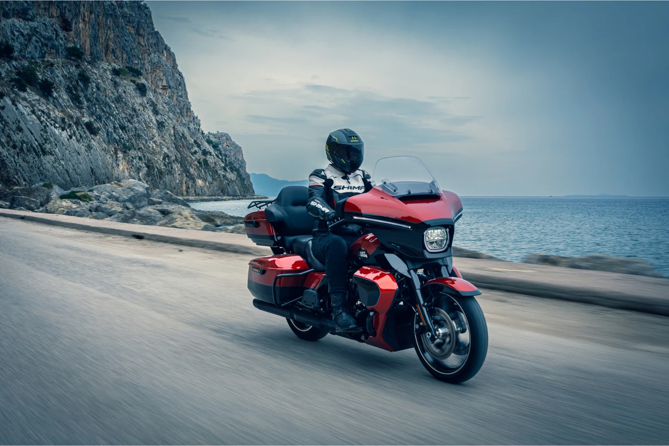 Czerwony motocykl typu cruiser prowadzony przez motocyklistę w czarnym kasku i skórzanej odzieży, jadący wzdłuż nadmorskiej, krętej drogi z wysokimi klifami po lewej stronie i spokojnym morzem po prawej.