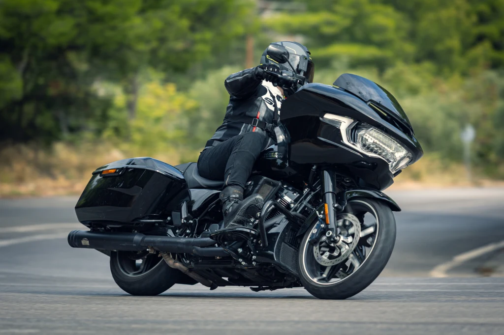 Motocyklista ubrany w czarny kombinezon i kask jedzie nowoczesnym czarnym motocyklem turystycznym po łuku asfaltowej drogi, w tle zielone drzewa.