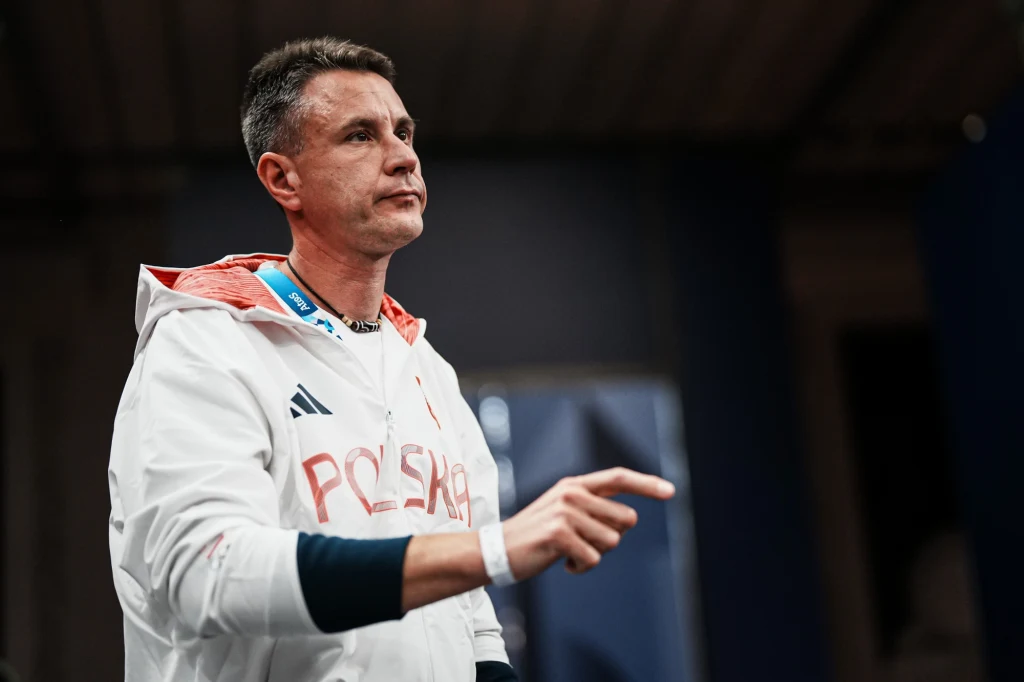 Bartłomiej Język Mężczyzna w sportowej kurtce z napisem Polska stoi w pomieszczeniu, wykonuje gest ręką wskazując palcem, ma na szyi identyfikator, otoczenie rozmyte i ciemne.