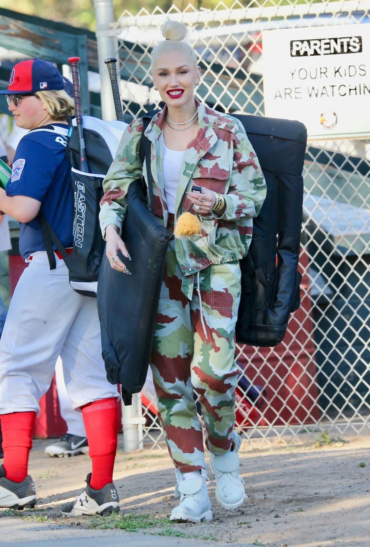 Kobieta w ubraniu w wojskowym stylu i białych sportowych butach trzyma duży czarny pokrowiec, uśmiechnięta na tle ogrodzenia boiska baseballowego, obok przechodzi chłopiec w stroju sportowym.