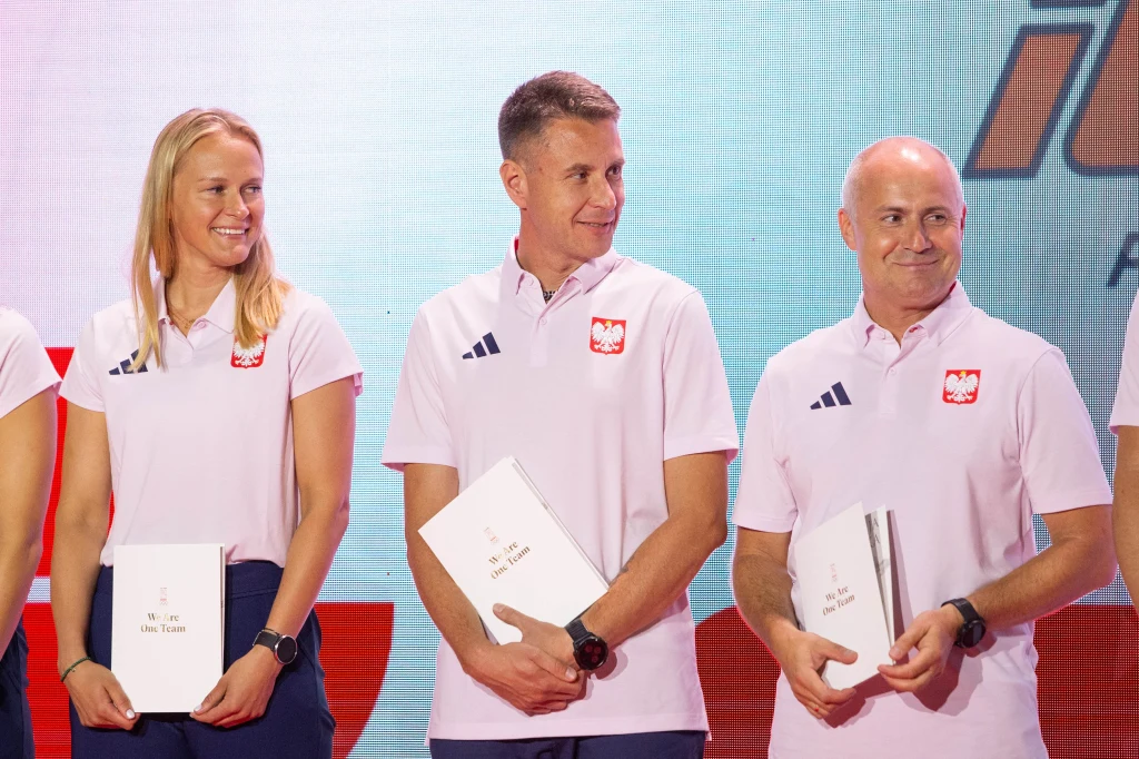 Od lewej: Hanna Łyczbińska, Bartłomiej Język i Artur Fajkis Trójka sportowców ubranych w białe koszulki z godłem Polski, trzymających w rękach dokumenty lub teczki, stojących razem i uśmiechających się na tle jasnej planszy z logotypem.