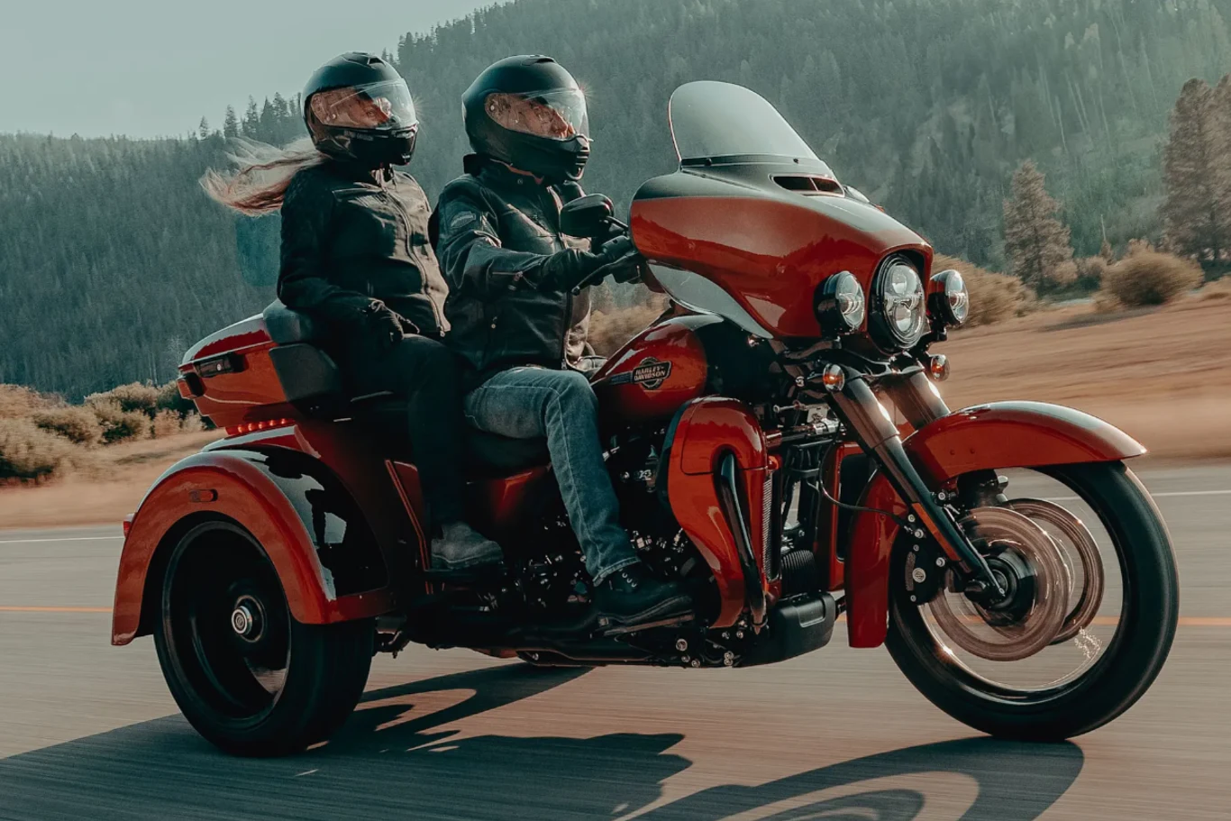 Dwie osoby w kaskach motocyklowych i ubraniach ochronnych jadą na czerwonym trójkołowym motocyklu po górskiej drodze, w tle widoczne są drzewa i wzgórza.