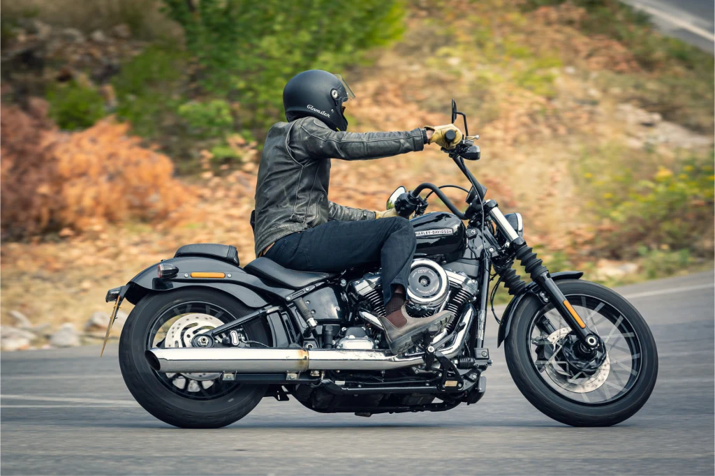 Motocyklista w czarnej skórzanej kurtce i kasku jedzie nowoczesnym motocyklem po zakręconej drodze, otoczonej naturalną roślinnością.