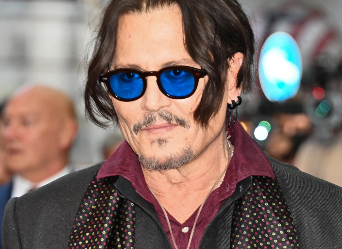 Johnny Depp założył własną wytwórnię Mężczyzna w okularach przeciwsłonecznych z niebieskimi szkłami, ubrany elegancko w ciemną marynarkę i wzorzysty szalik, z zarostem i dłuższymi, częściowo zaczesanymi na bok włosami.