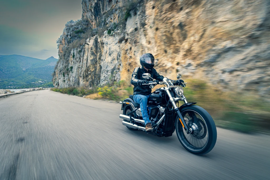 Motocyklista w kasku i skórzanej kurtce jedzie klasycznym motocyklem po krętej, górskiej drodze, obok skalistego urwiska, w tle widać zieloną dolinę i wzgórza.