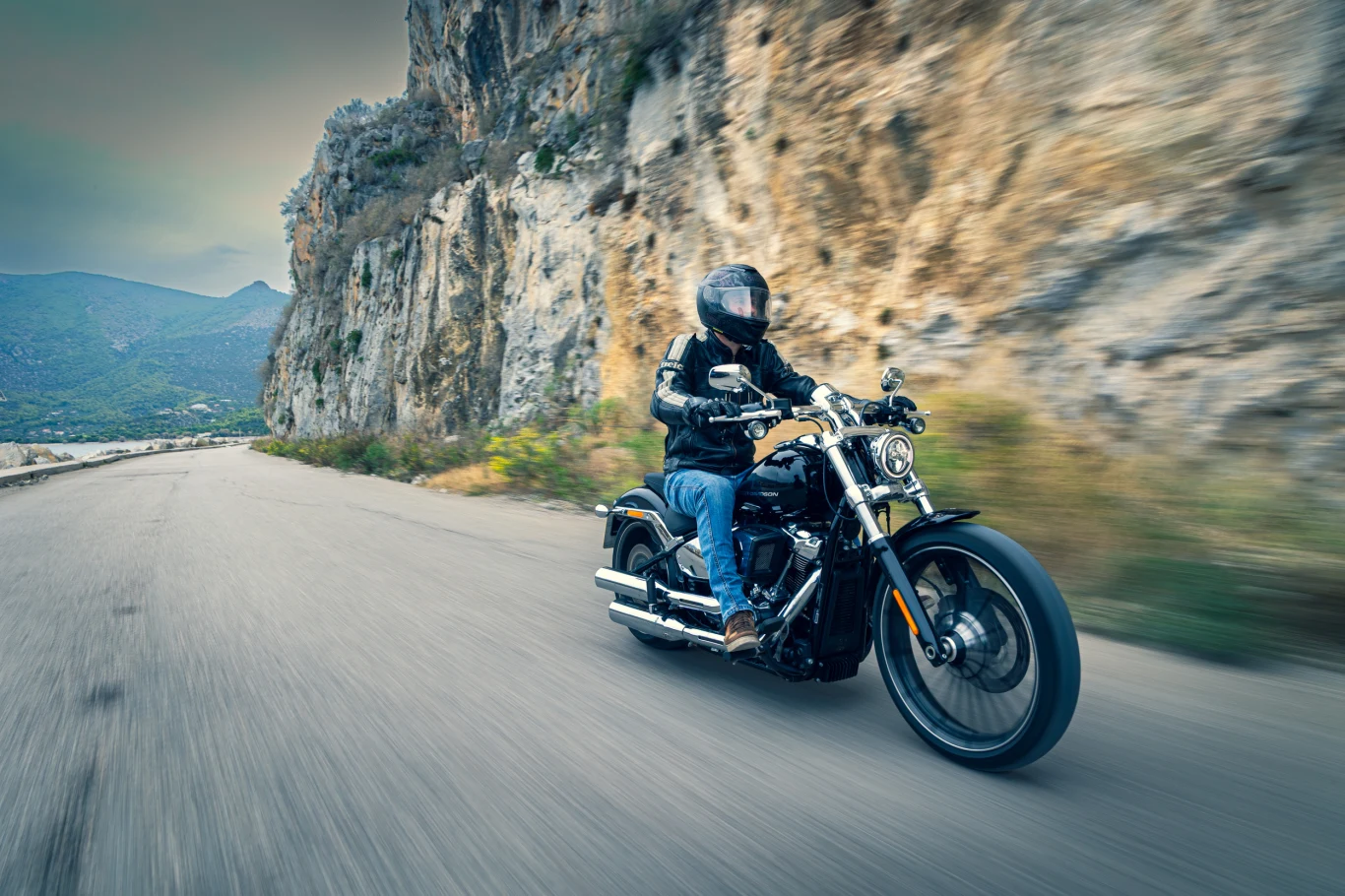 Motocyklista w kasku i skórzanej kurtce jedzie klasycznym motocyklem po krętej, górskiej drodze, obok skalistego urwiska, w tle widać zieloną dolinę i wzgórza.