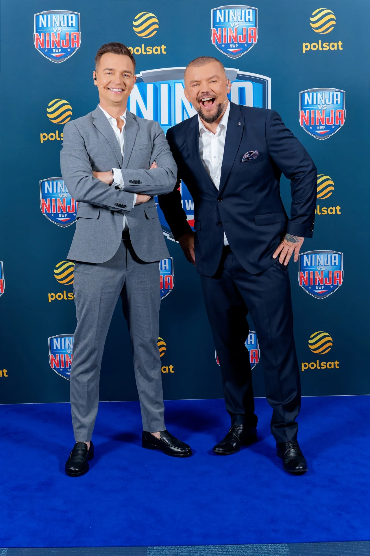 Dwóch elegancko ubranych mężczyzn w garniturach stoi na niebieskim dywanie na tle ściany z logotypami programu Ninja Warrior Poland i kanału Polsat, obaj uśmiechnięci, pozujący w przyjaznej atmosferze.