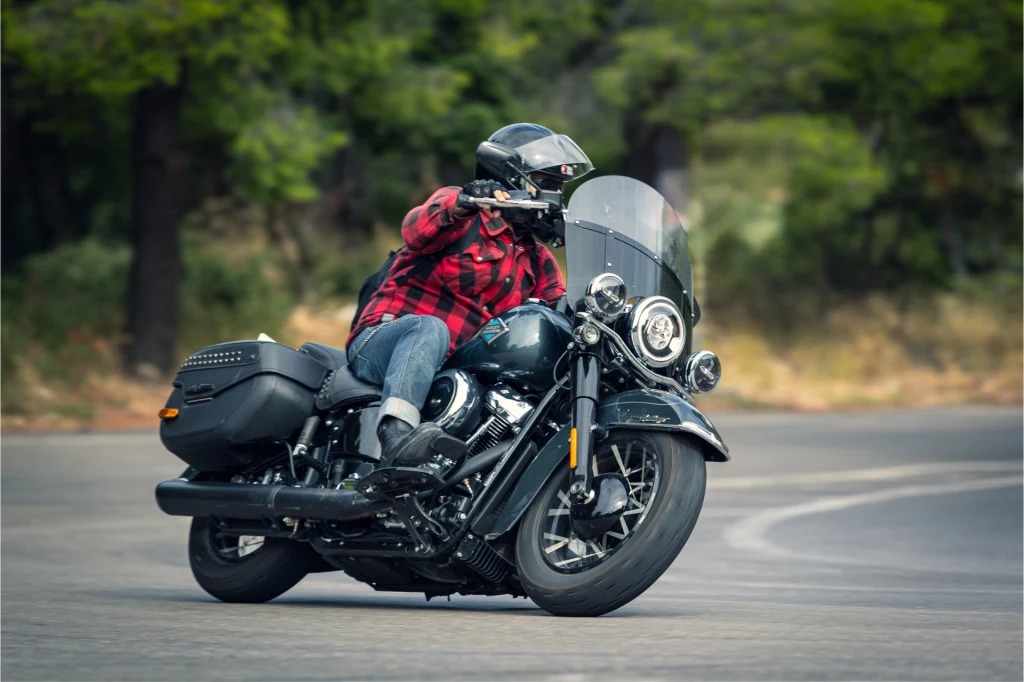 Motocyklista w kasku i czerwonej koszuli flanelowej pochylający się podczas jazdy czarnym motocyklem po zakręcie, w tle rozmyte drzewa i asfaltowa droga.