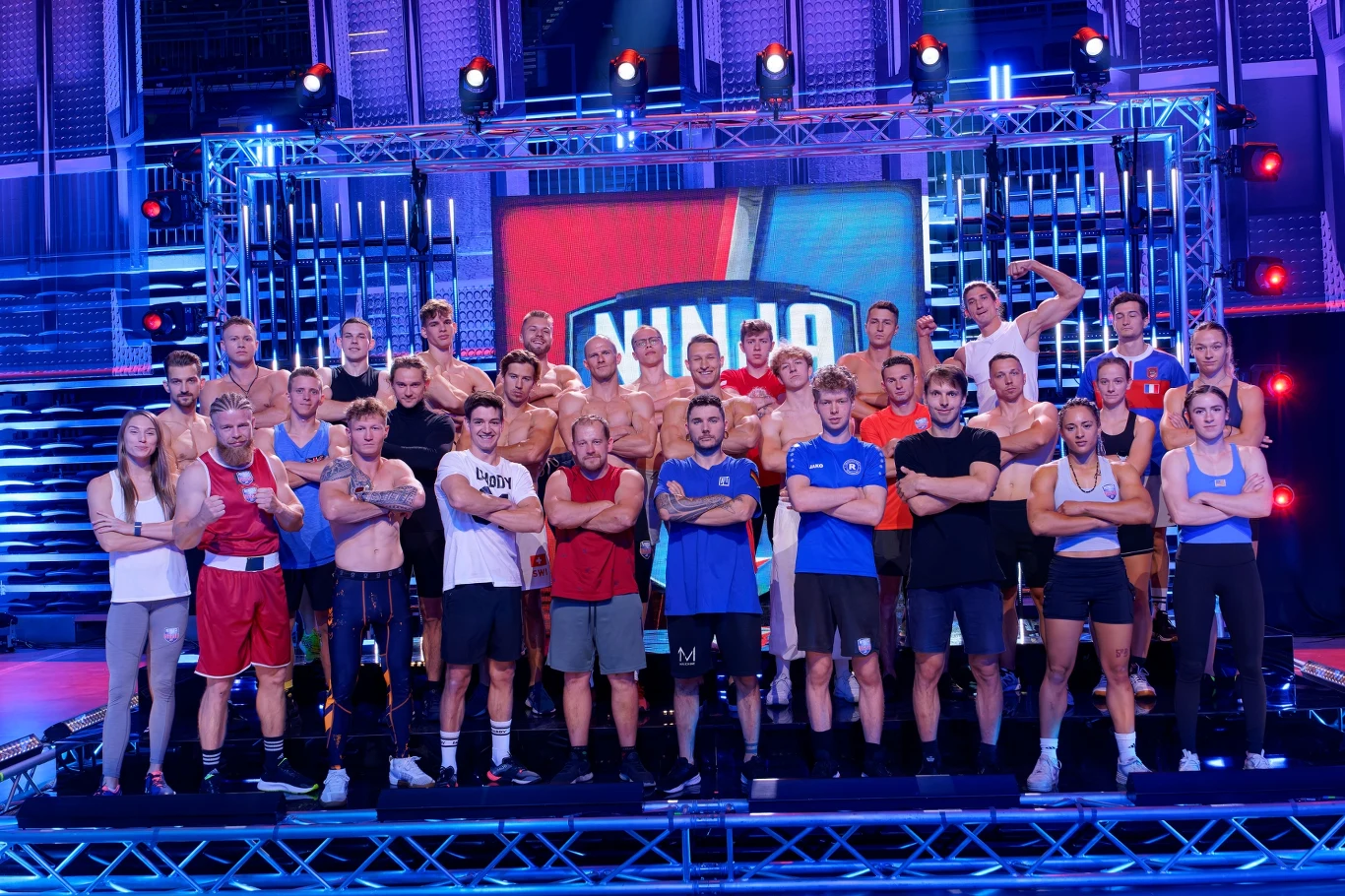 Ninja vs Ninja Grupa kobiet i mężczyzn w sportowych strojach stoi na scenie, w tle duży ekran z logiem programu rozrywkowego, oświetlenie sceniczne podkreśla dynamiczny charakter wydarzenia.