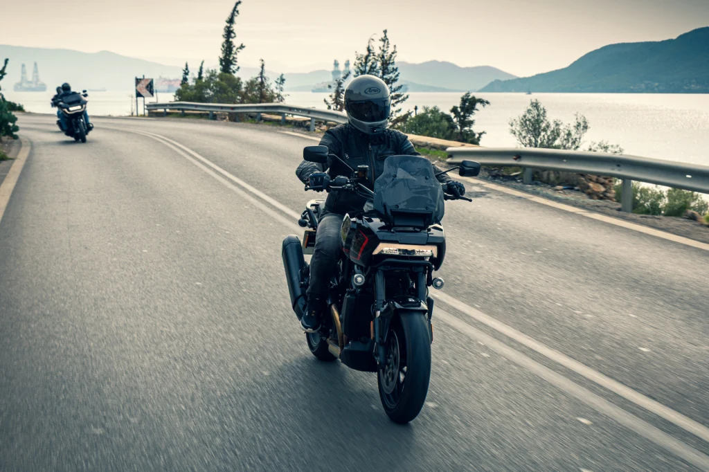 Dwóch motocyklistów jadących szeroką, asfaltową drogą nad wodą, otoczoną górami i roślinnością, z kaskami ochronnymi i ubraniami motocyklowymi, przy lekko pochmurnym niebie.