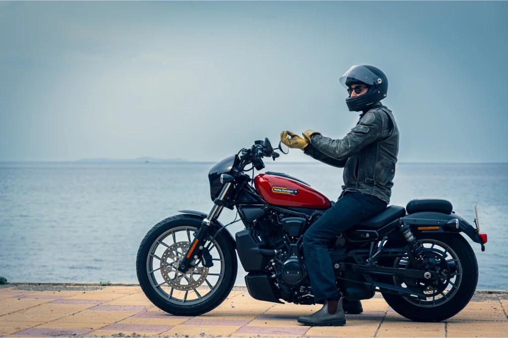 Mężczyzna w czarnym kasku, skórzanej kurtce i rękawiczkach siedzi na czerwonym motocyklu zaparkowanym przy nabrzeżu z widokiem na spokojną wodę.