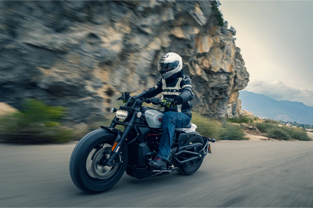 Motocyklista w pełnym stroju ochronnym oraz kasku porusza się nowoczesnym motocyklem po krętej asfaltowej drodze w górskim terenie, w tle widać wysokie skały i rozległe widoki na dolinę.