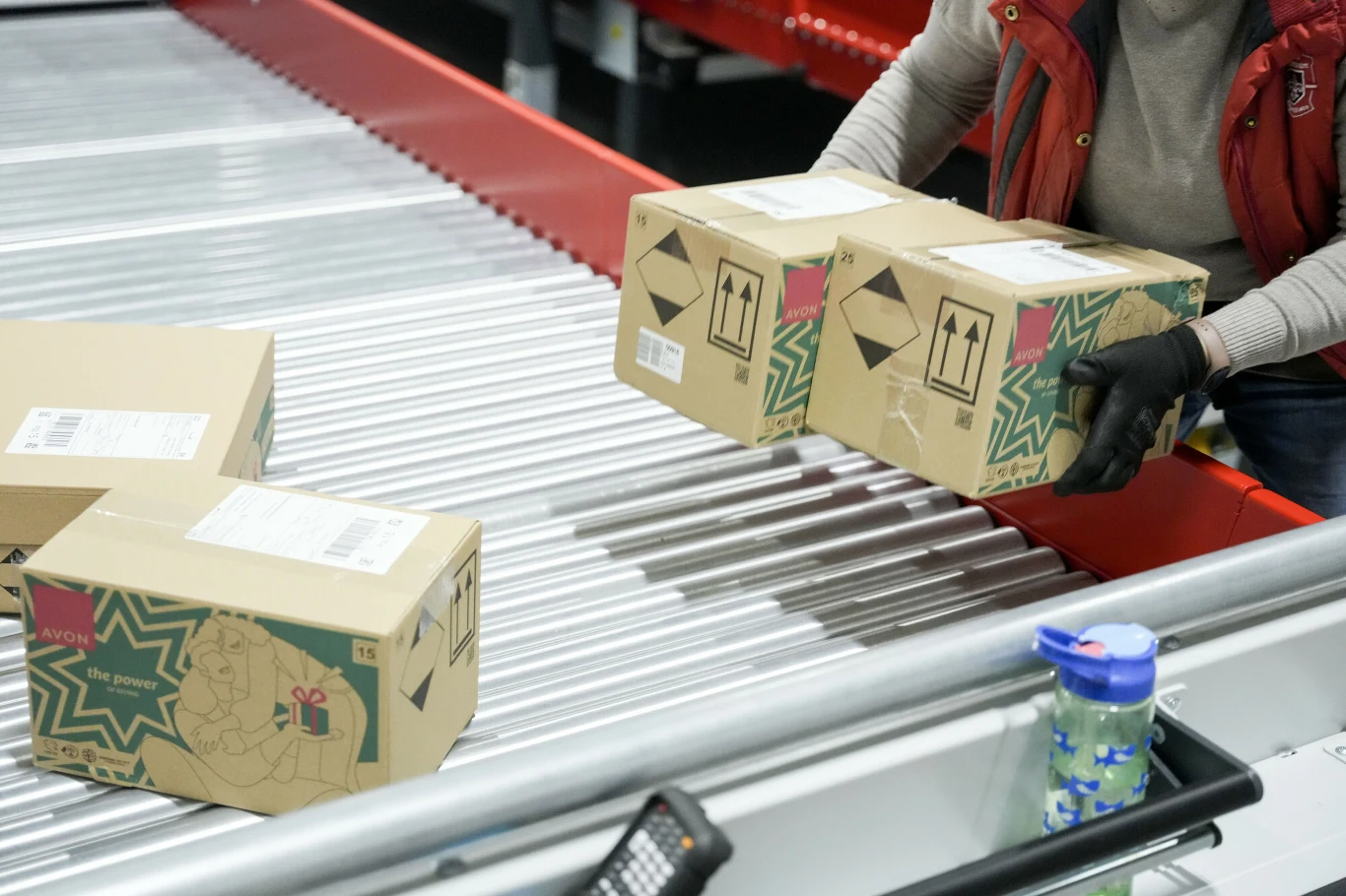 Miliony paczek do dostarczenia. Kurierzy na pełnych obrotach, by zdążyć przed świętami (zdj, ilustracyjne) Ręce osoby w rękawiczkach układającej kartonowe paczki na taśmie transportowej w centrum logistycznym, w tle widoczna woda w butelce oraz fragment urządzenia do skanowania.