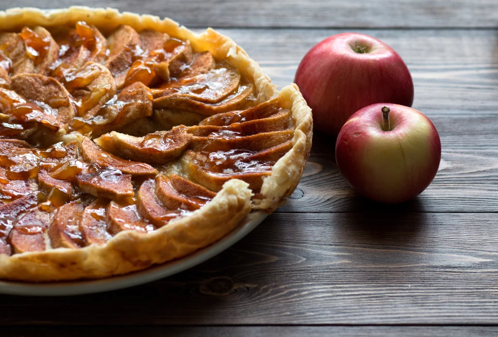 Najlepszą odmianą jabłek na ciasto będzie Antonówka, Szara Reneta lub Granny Smith Szarlotka z plasterkami jabłek na kruchym cieście podana na białym talerzu, obok dwa świeże czerwone jabłka na drewnianym blacie.
