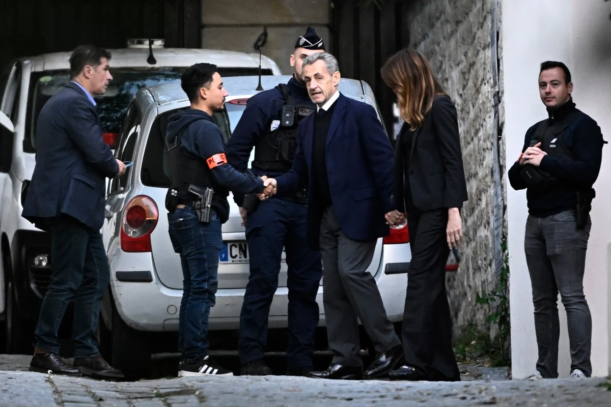 Nicolas Sarkozy tuż przed zamknięciem go w więzieniu Grupa pięciu osób zgromadzona w pobliżu zaparkowanego białego samochodu, dwaj mężczyźni, z których jeden ubrany jest w mundur policjanta, wymieniają uścisk dłoni; po prawej stronie kobieta trzyma mężczyznę za rękę, a obok nich stoi jeszcze dwóch mężczyzn.