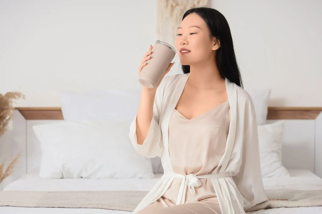 Lepszy sen, mniej uderzeń gorąca, łagodniejsze objawy menopauzy Młoda kobieta siedzi na łóżku w jasnym pokoju, ubrana w beżową piżamę i biały szlafrok, trzyma w dłoni kubek termiczny i delektuje się napojem.