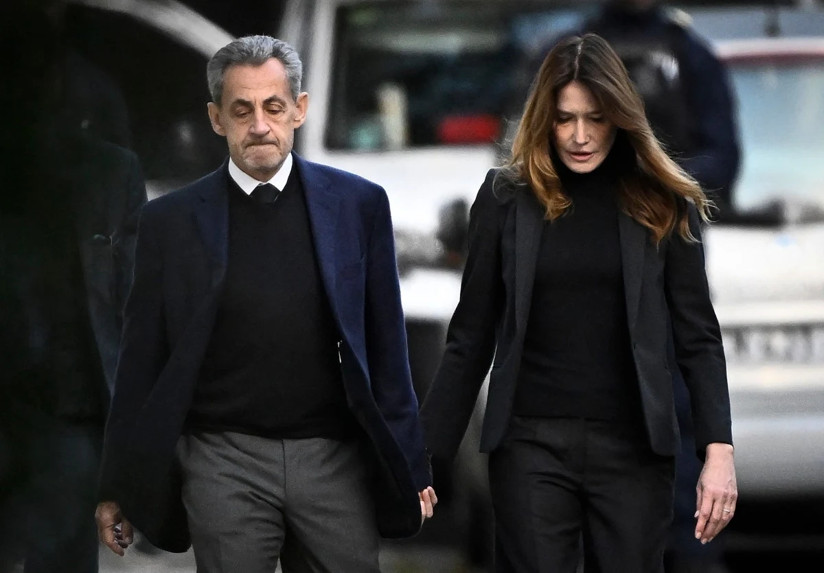 Nicolas Sarkozy z Carlą Bruni w drodze do więzienia Dwoje elegancko ubranych dorosłych idzie razem, trzymając się za ręce, w tle widoczny pojazd oraz niewyraźne sylwetki osób.