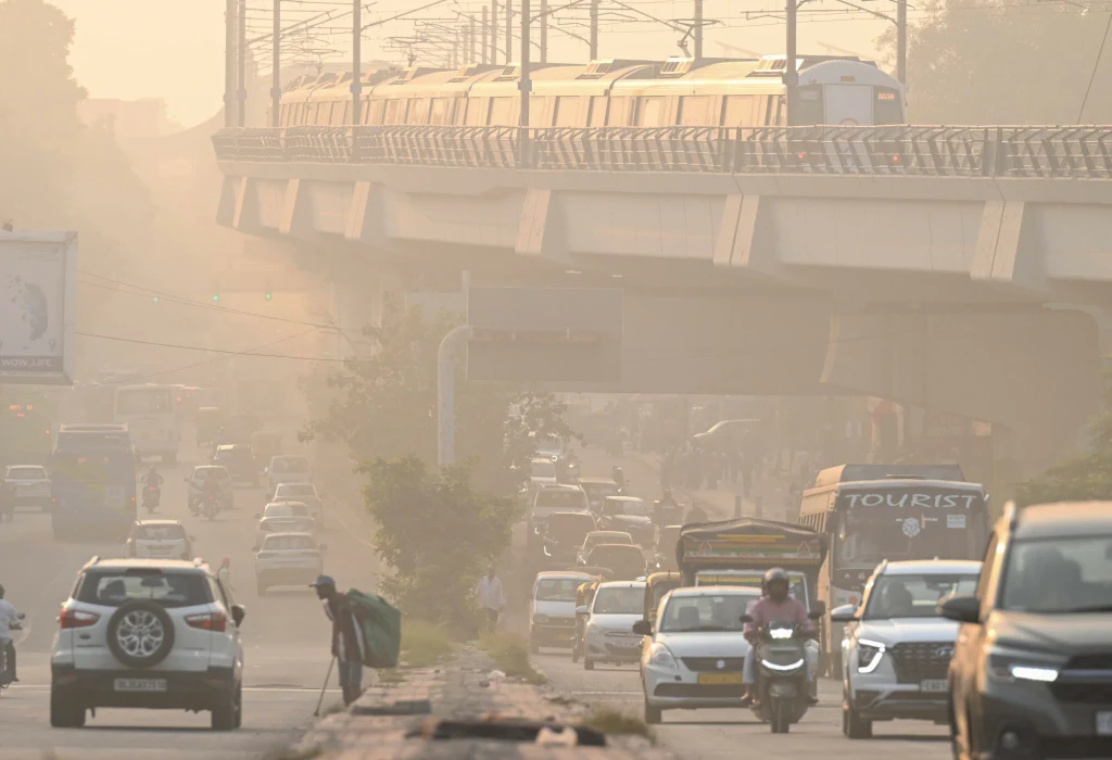 Gęsty ruch uliczny pod estakadą metra otoczony mgłą lub smogiem, pieszy schylający się na poboczu, liczne samochody oraz motocykl, słaba widoczność wskutek zanieczyszczenia powietrza.