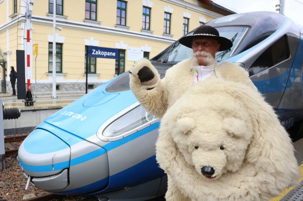 Biały miś przywitał Pendolino w Zakopanem Mężczyzna ubrany w tradycyjny strój góralski oraz kostium przypominający niedźwiedzia polarnego stoi na peronie przy nowoczesnym pociągu, w tle widoczna tablica z napisem Zakopane oraz charakterystyczny budynek dworca.