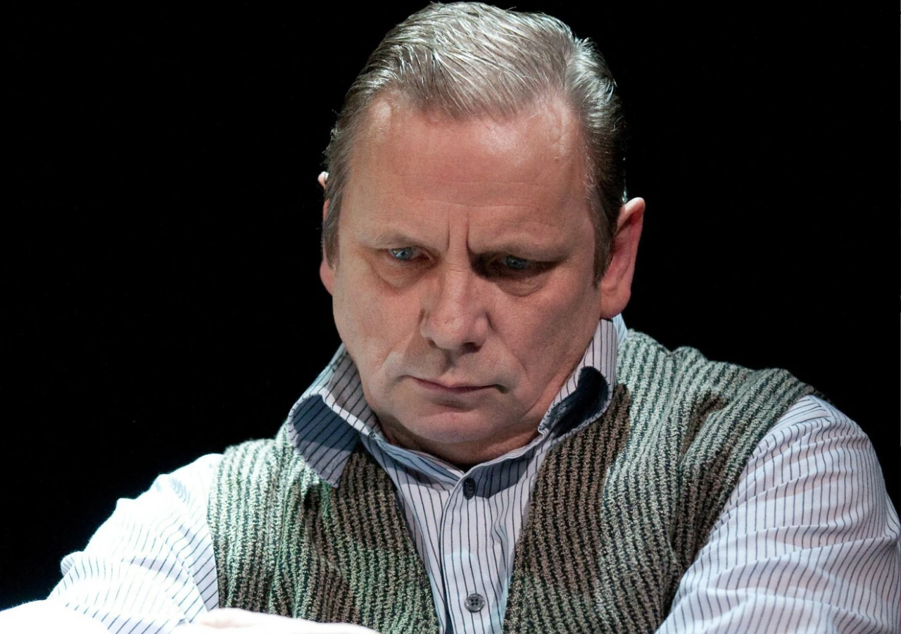 Mężczyzna o poważnym wyrazie twarzy, ubrany w sweter bez rękawów na koszuli w pionowe pasy, na ciemnym tle.