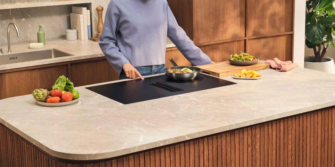 Uciążliwe zabrudzenia na płycie? Wybierz płytę indukcyjną SaphirMatt® z trwałą, odporną i łatwą w czyszczeniu powierzchnią Osoba stojąca za nowoczesnym blatem kuchennym przygotowuje potrawy, na blacie widoczne talerze z warzywami, gotowane danie na patelni oraz przekąski, w tle drewniane szafki i umywalka.