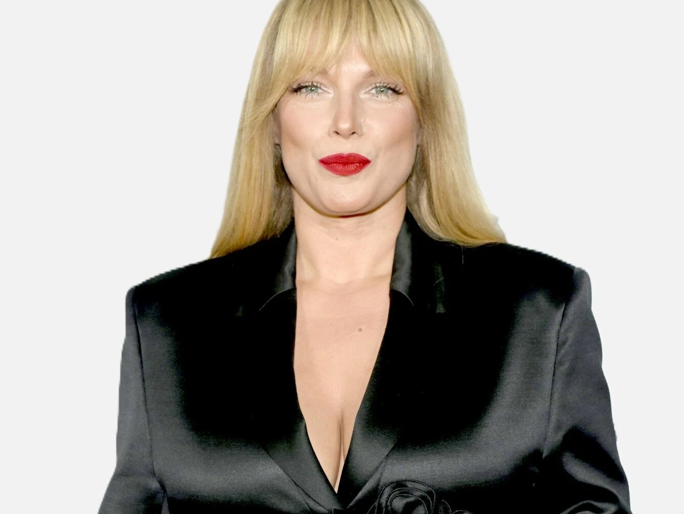 Joanna Liszowska przeszła zaskakującą metamorfozę Kobieta o prostych blond włosach i prostych grzywkach, ubrana w czarną, satynową marynarkę z głębokim dekoltem, patrzy prosto w obiektyw aparatu, ma intensywny makijaż, podkreślone czerwone usta, jasne tło.