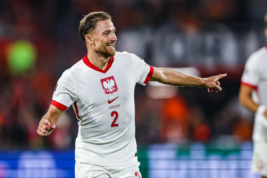 Polski piłkarz w białej koszulce z numerem 2 i godłem Polski na piersi biegnie po boisku, uśmiecha się i wskazuje ręką w bok, w tle rozmyty widok trybun i innych zawodników.