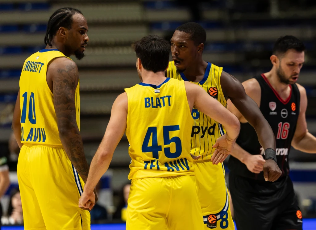 Koszykarze w żółtych strojach drużyny Maccabi Tel Aviv omawiają taktykę podczas meczu, w tle widoczny zawodnik drużyny przeciwnej w czarnym stroju.