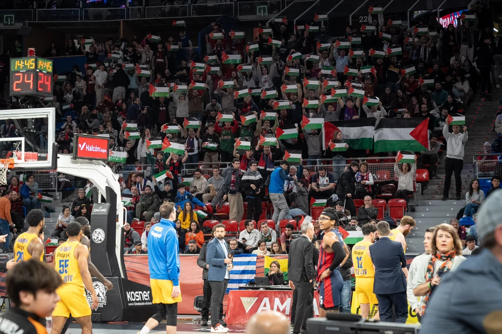 Koszykarskie rozgrywki z udziałem zawodników w żółtych i niebieskich strojach na tle trybun, gdzie tłum kibiców trzyma palestyńskie flagi i transparenty, wyrażając swoje poparcie w czasie meczu.