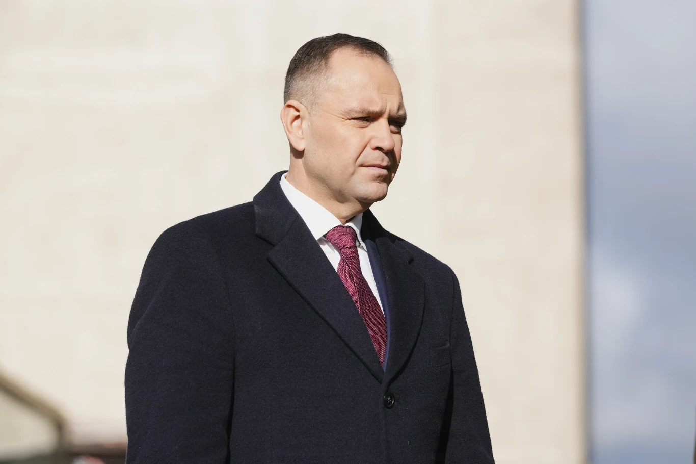 Karol Nawrocki był szefem IPN w latach 2021-2025 Mężczyzna w średnim wieku w eleganckim płaszczu i bordowym krawacie stoi na zewnątrz na tle jasnego budynku, spoglądając w bok z poważnym wyrazem twarzy.