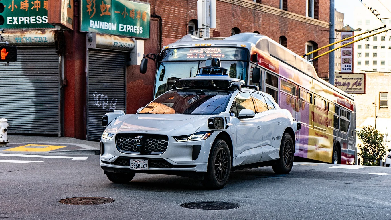 Taksówka Waymo na ulicach San Francisco Biały samochód autonomiczny wyposażony w czujniki i kamery porusza się miejską ulicą, w tle kolorowy autobus oraz budynki z azjatyckimi szyldami.