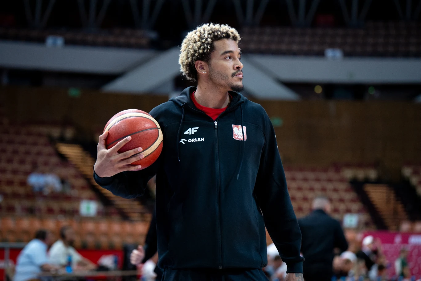 Młody mężczyzna w sportowej czarnej bluzie z logotypami trzyma piłkę do koszykówki na hali sportowej, w tle rozmazane trybuny i inne osoby.