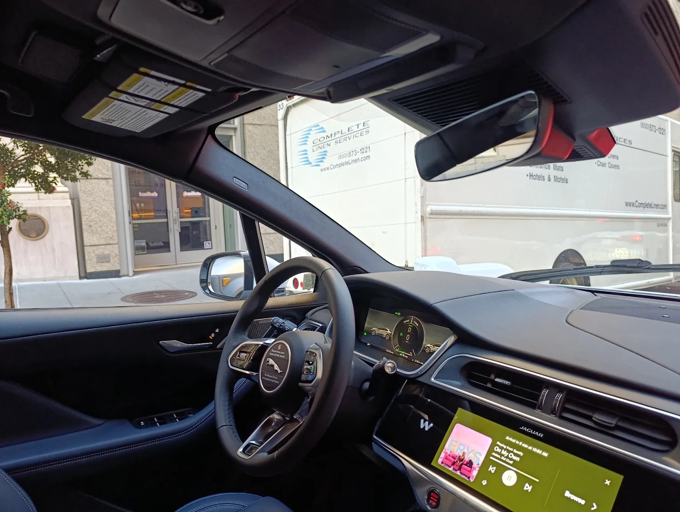 Jazda samochodem bez kierowcy na pewno robi surrealistyczne wrażenie Wnętrze nowoczesnego samochodu z cyfrowym panelem sterowania, dużym ekranem dotykowym wyświetlającym muzykę i kierownicą z logo Jaguara, przez okno widać fragment białej furgonetki z logotypem firmy.