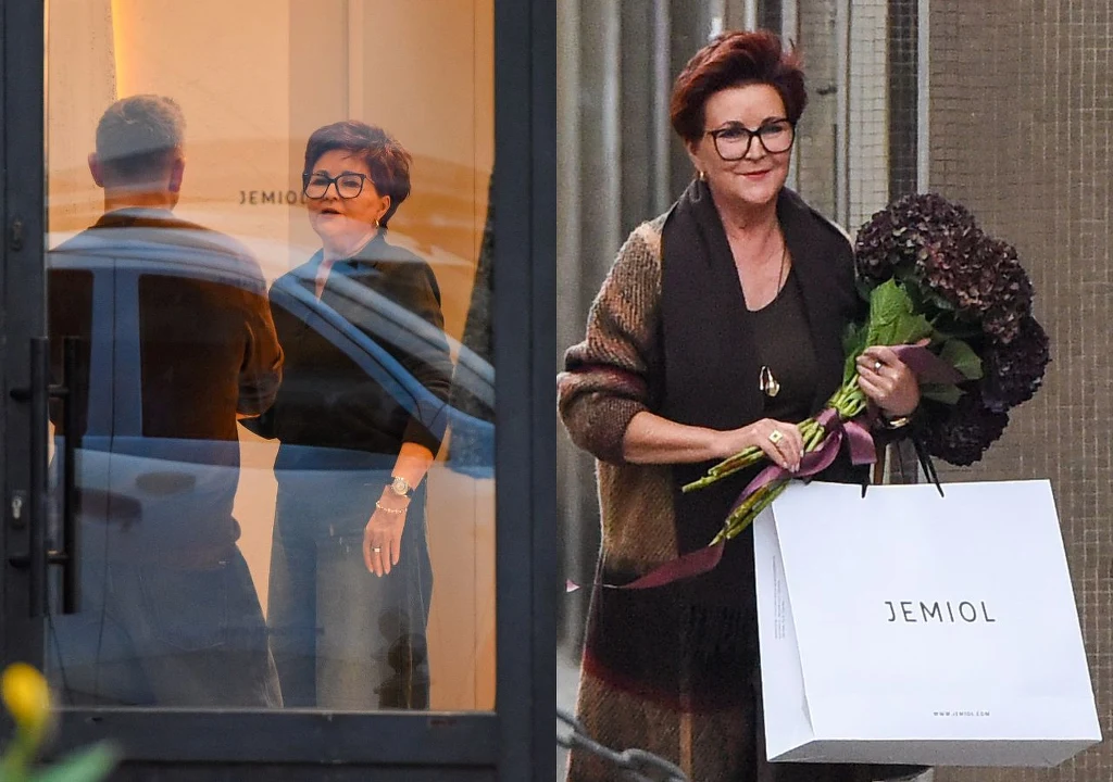 Jolanta Kwaśniewska na zakupach w butiku Łukasza Jemioła Elegancka kobieta z krótkimi ciemnymi włosami i okularami trzyma bukiet ciemnych kwiatów oraz białą torbę zakupową z napisem JEMIOL, uśmiecha się i opuszcza butik.