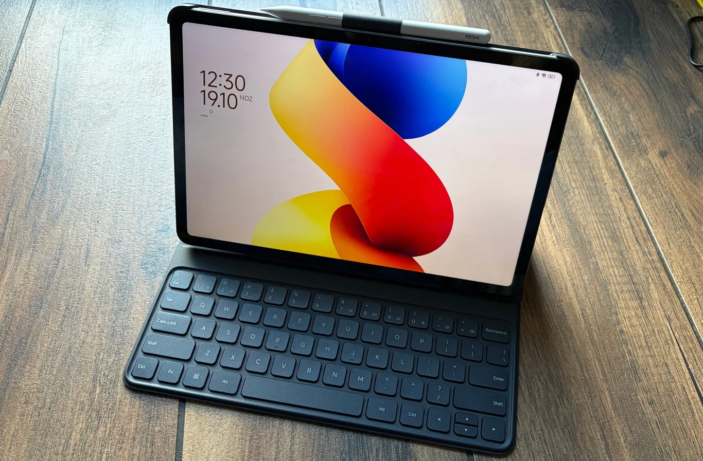 Test Redmi Pad 2 Pro. Niedrogi tablet, który pozytywnie zaskakuje. Tablet z podłączoną klawiaturą oraz rysikiem umieszczonym na górnej krawędzi ekranu, całość ustawiona na drewnianym blacie, na ekranie kolorowa tapeta oraz widoczne aktualne godzina i data.