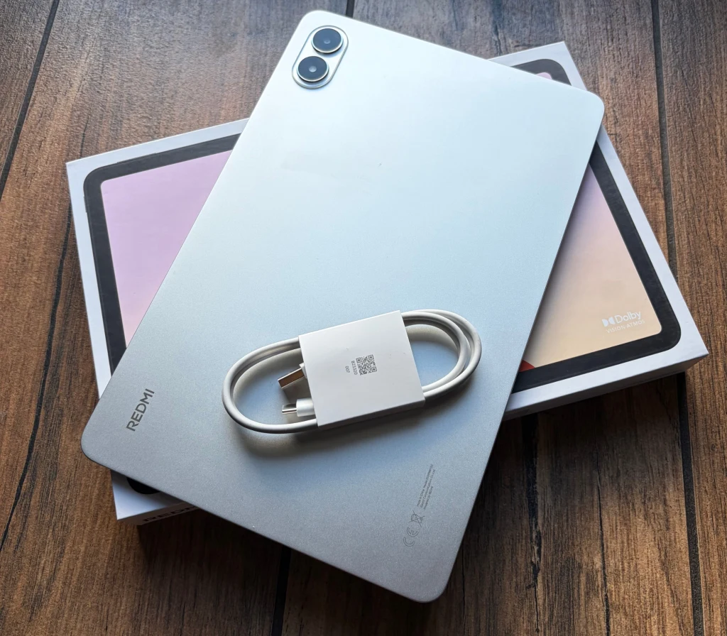 Zawartość opakowania z Redmi Pad 2 Pro. Srebrny tablet marki Redmi położony na pudełku, na nim umieszczony zwinięty kabel USB typu C, całość na drewnianym blacie.