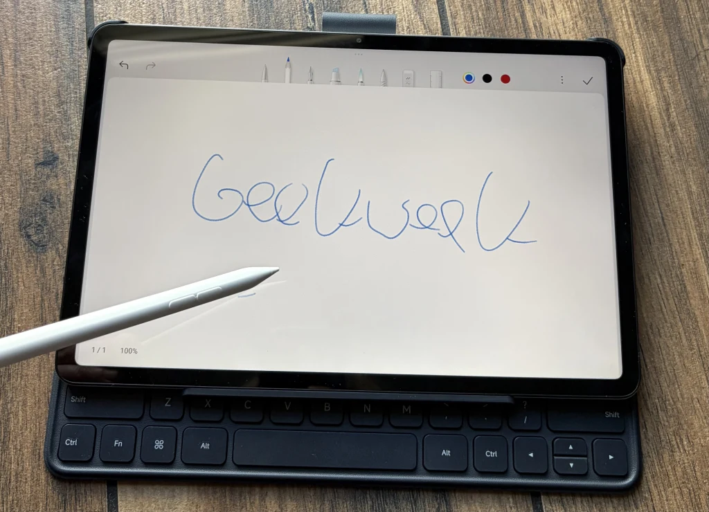 Redmi Pad 2 Pro pozwala na pracę z użyciem rysika Smart Pen. Tablet z klawiaturą oraz rysikiem, na ekranie widoczny odręczny napis 'Geekweek', urządzenie leży na drewnianym blacie.