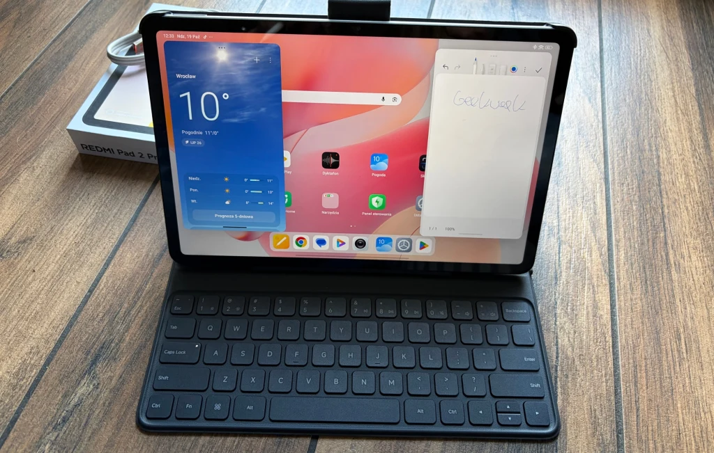 Redmi Pad 2 Pro pozwala na pracę z aplikacjami w pływających oknach. Tablet z podłączoną klawiaturą wyświetla ekran główny z pogodą i notatką, stoi na drewnianym blacie. W tle widoczne opakowanie urządzenia.