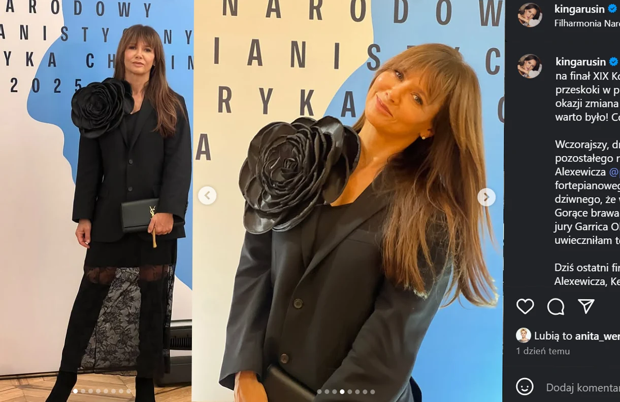 Kobieta w eleganckiej, czarnej marynarce z dużym, ozdobnym kwiatem na ramieniu, trzymająca kopertówkę, stoi na tle ściany z napisem związanym z wydarzeniem muzycznym. Długie, rozpuszczone włosy, czarna spódnica z koronką dopełniają stylizację.