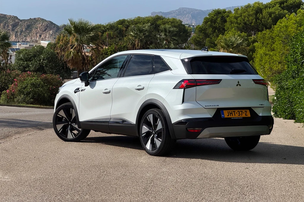Biały samochód Mitsubishi ASX zaparkowany na asfaltowej drodze w otoczeniu zielonych drzew i górskich krajobrazów, widoczny z tyłu z holenderską rejestracją oraz sportowym wyglądem.