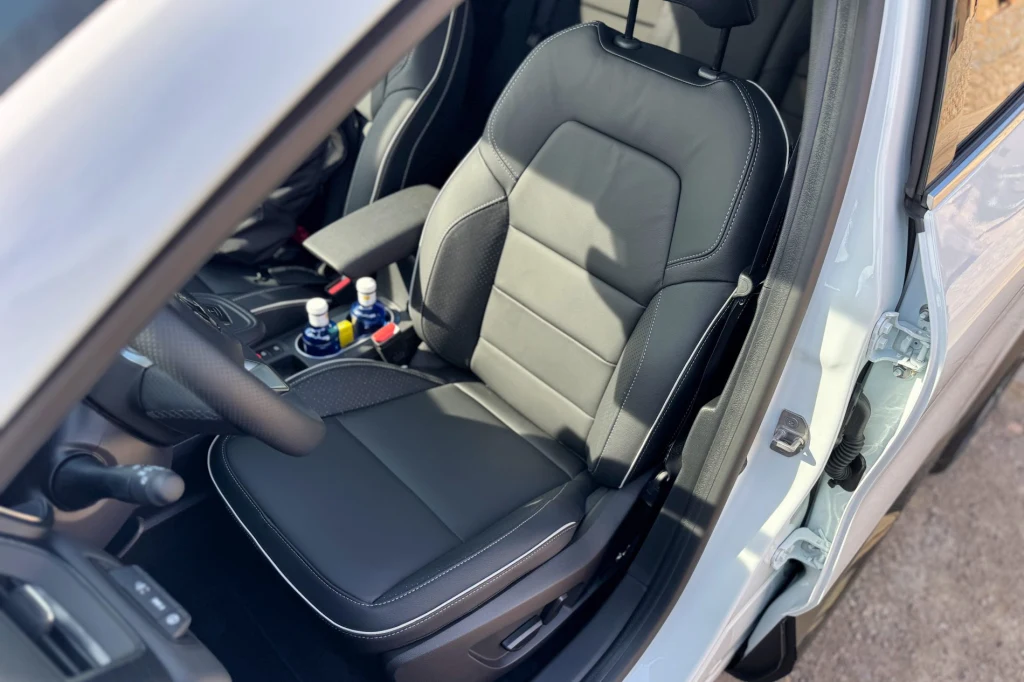 Czarne, skórzane siedzenie kierowcy w samochodzie z widocznym podłokietnikiem oraz konsolą środkową, na której znajdują się butelki z napojami i uchwyty.