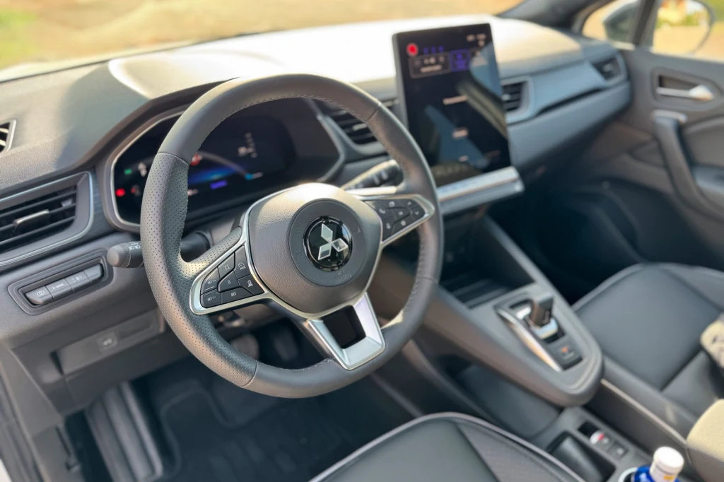 Wnętrze nowoczesnego samochodu z widoczną kierownicą, cyfrowym panelem wskaźników oraz dużym dotykowym ekranem na konsoli centralnej, fotele i detale wnętrza wykończone w ciemnych tonacjach.