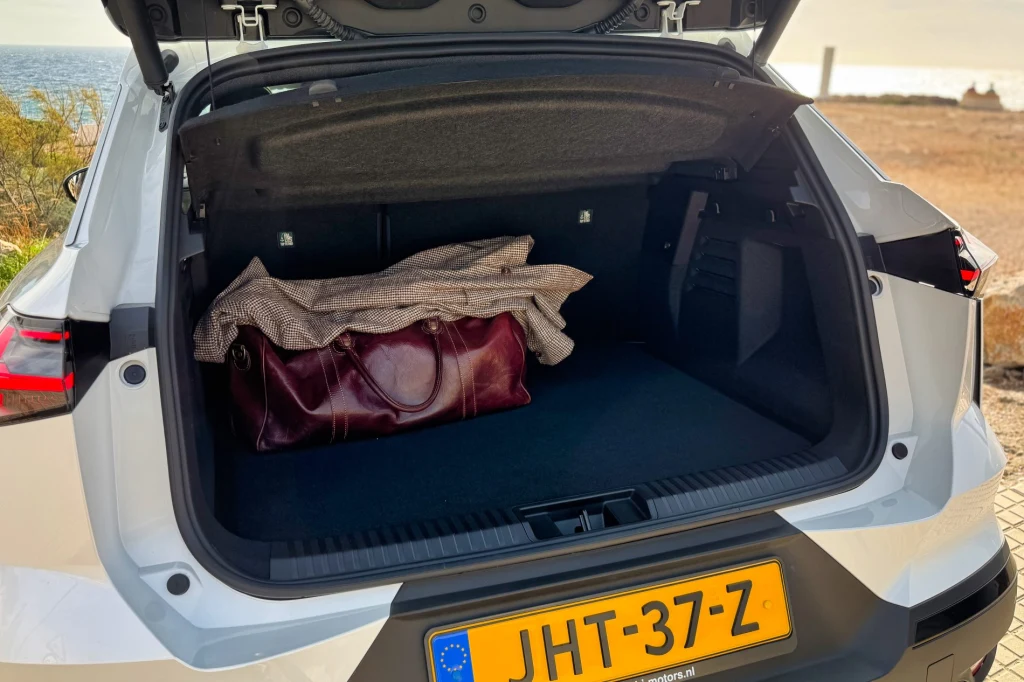 Otwarty bagażnik samochodu z żółtymi tablicami rejestracyjnymi, w środku leży torba podróżna przykryta swetrem, wokół teren pozamiejski z suchą roślinnością.