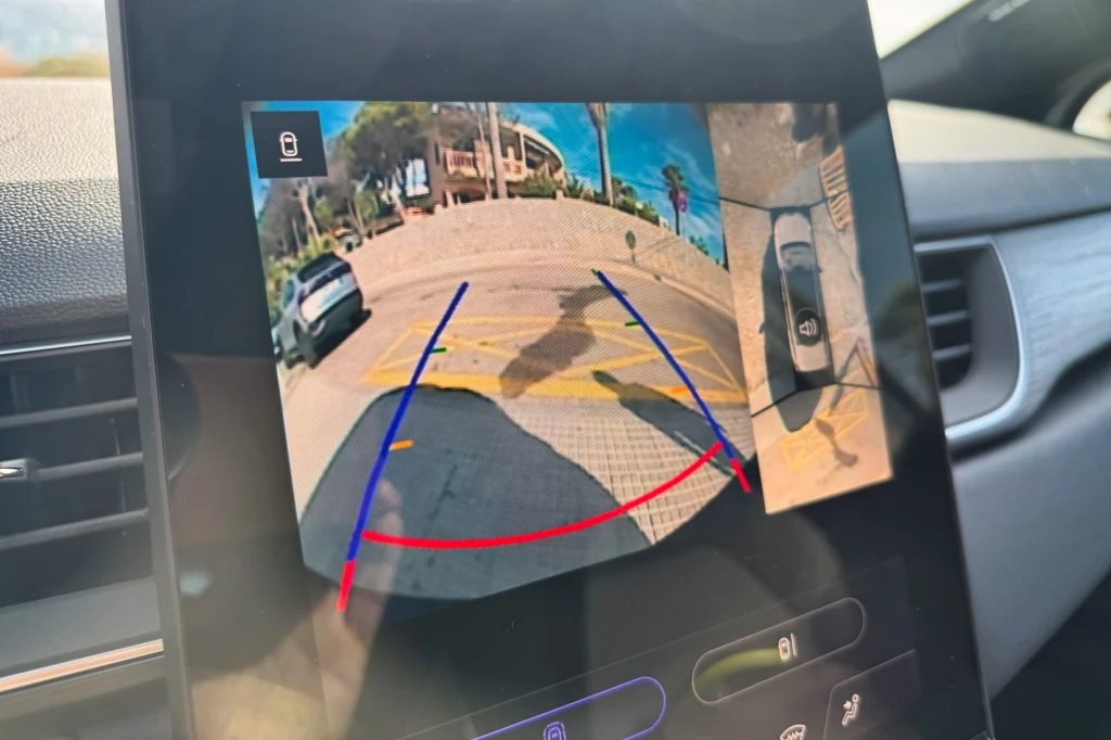 Widok ekranu samochodowego z włączoną kamerą cofania, na której widać fragment drogi, zaparkowany samochód po lewej stronie oraz kolorowe linie pomocnicze ułatwiające manewrowanie.