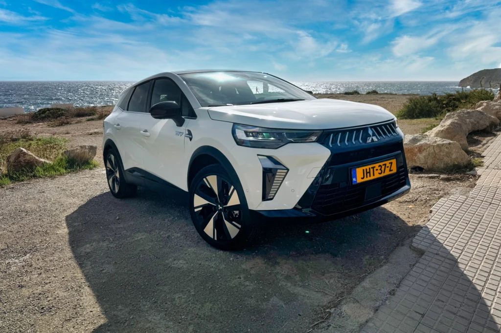 Biały samochód Mitsubishi z holenderską rejestracją zaparkowany na wybrzeżu, w tle widać morze, niebieskie niebo z lekkim zachmurzeniem oraz skaliste otoczenie.