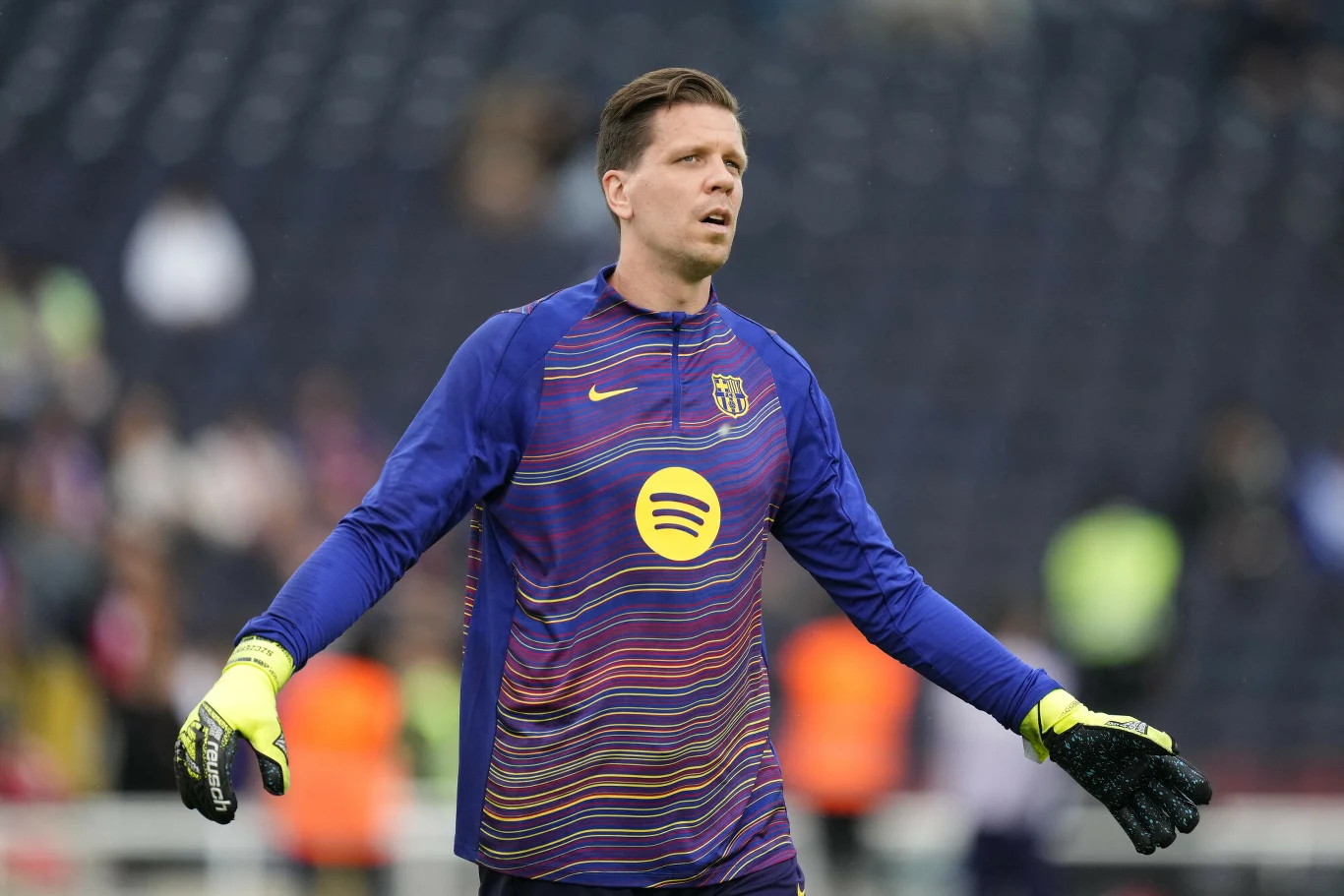 Wojciech Szczęsny Bramkarz w sportowym stroju treningowym klubu FC Barcelona na boisku, z rękami lekko uniesionymi, w tle niewyraźna trybuna stadionu.